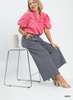 Woman wearing a pink blouse and gray pants sitting on a white stool against a white background.