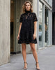 A woman modeling a short-sleeve black lace dress with a high mock neck and a flared skirt, standing on a city sidewalk next to a concrete building.