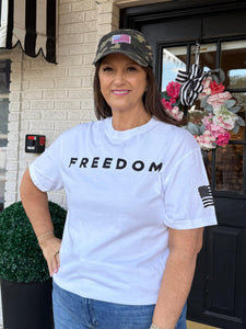 Woman wearing a white t-shirt with 'FREEDOM' printed on it, standing in front of a house entrance.